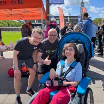 Zwei Männer stehen neben einer Frau, die in einem Rollstuhl sitzt, alle lächeln in die Kamera.