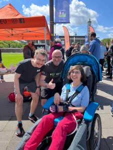 Zwei Männer stehen neben einer Frau, die in einem Rollstuhl sitzt, alle lächeln in die Kamera.