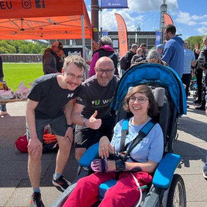 Zwei Männer stehen neben einer Frau, die in einem Rollstuhl sitzt, alle lächeln in die Kamera.