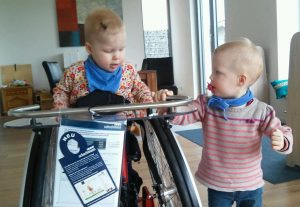Zwei kleine Kinder spielen an einem Kinderrollstuhl. Eines sitzt im Rollstuhl eines steht daneben.